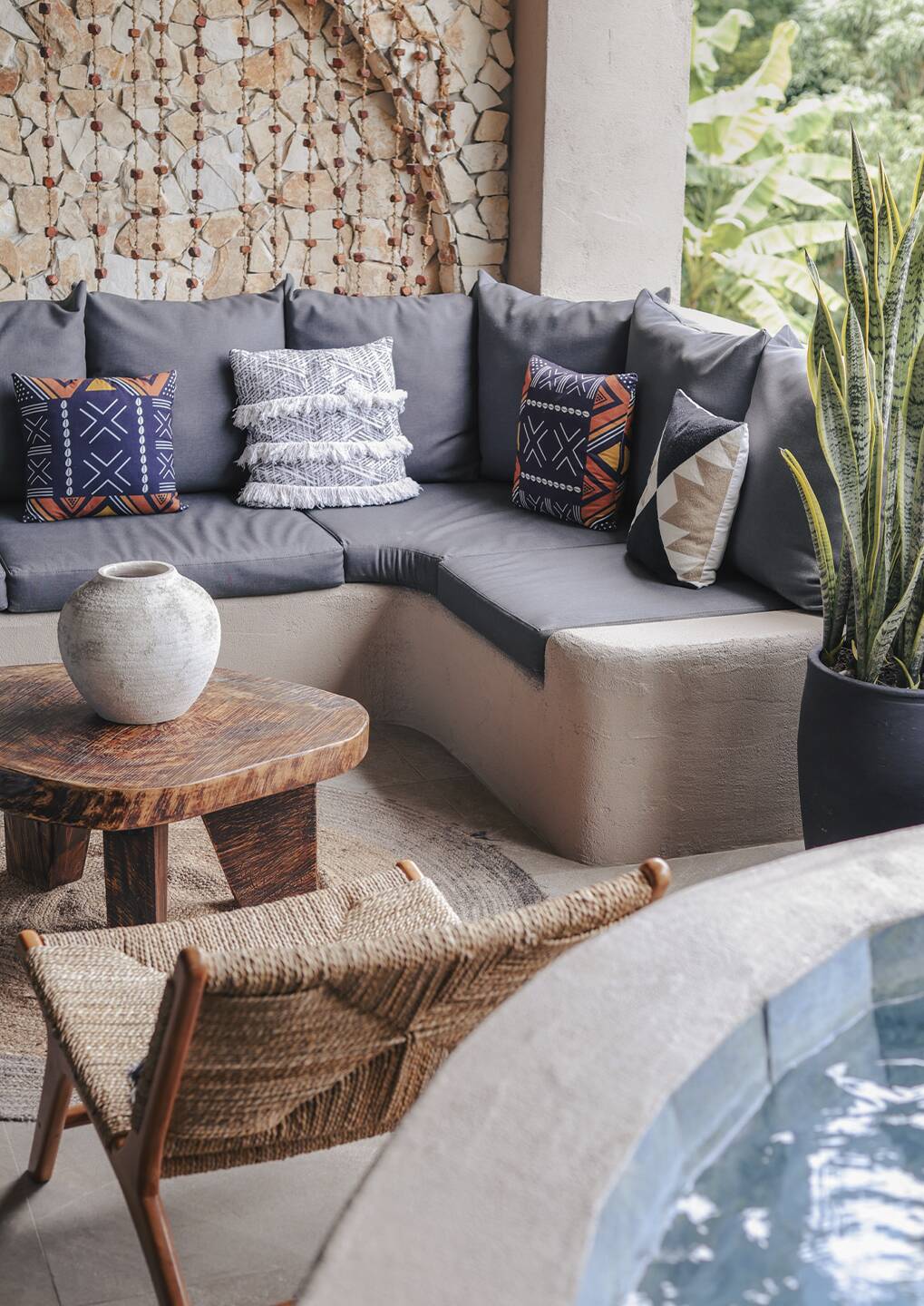 Curved gray outdoor sectional with patterned cushions, a round wooden table with a ceramic vase, a woven chair, and a potted plant by the pool.