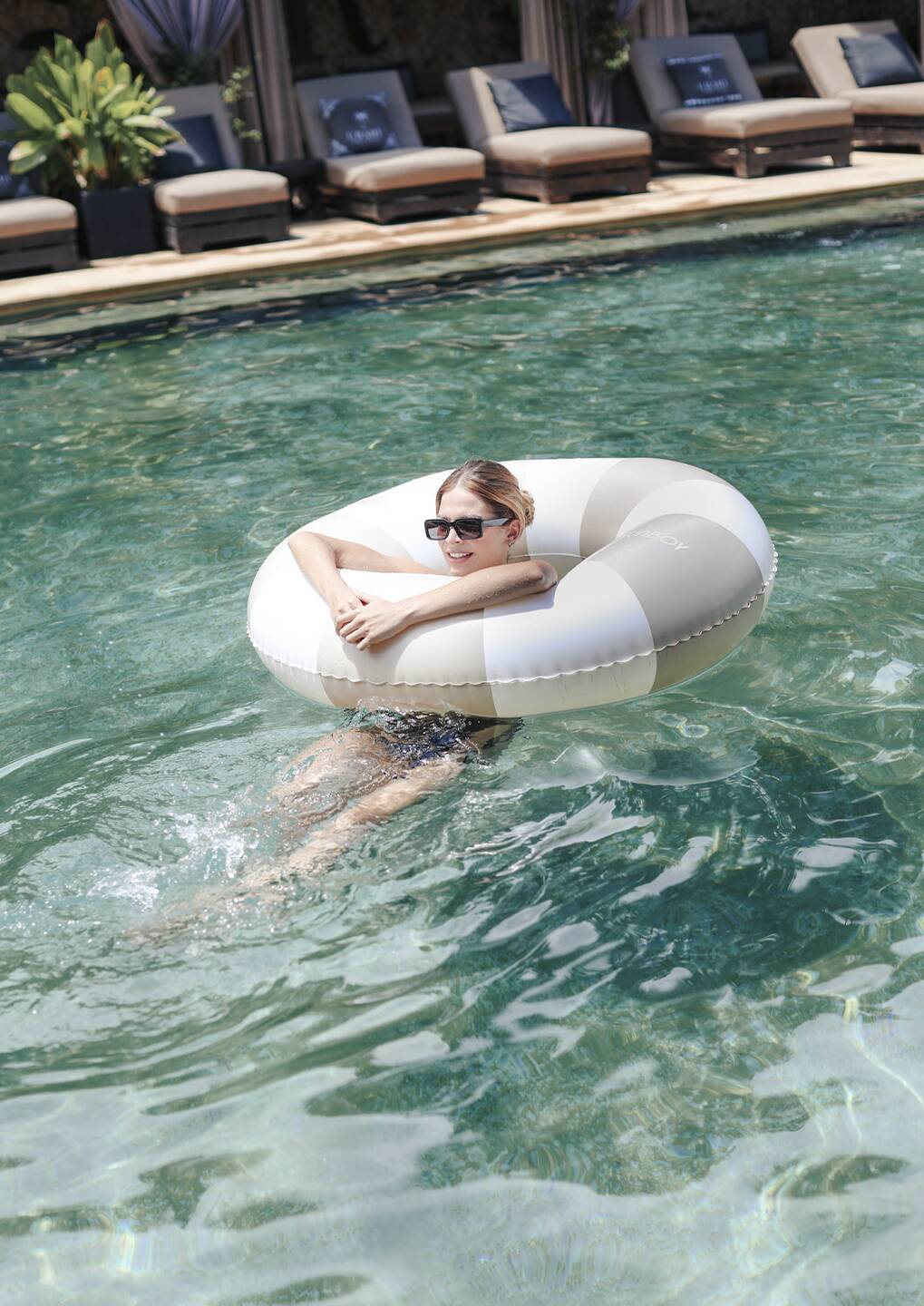 Woman wearing sunglasses relaxing in a striped inflatable pool ring, floating in a clear turquoise pool with resort lounge chairs in the background.