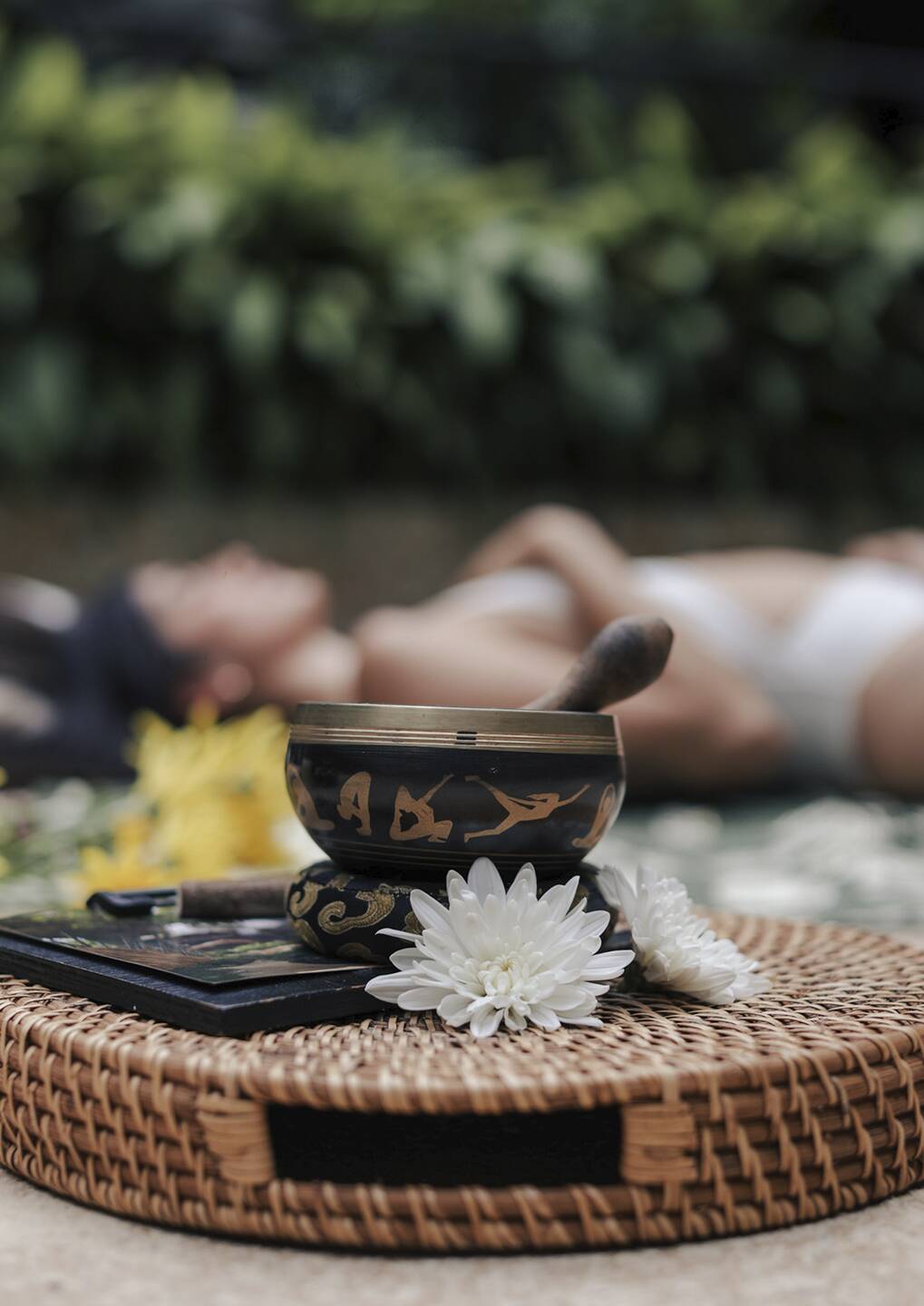Decorative singing bowl with white flowers on a woven mat, a person relaxing blurred in the background.