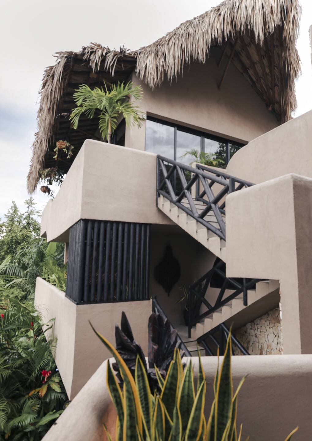 B4 4 - Casa Candela - Casa Candela Two-story tropical-style building with a thatched roof, beige stucco walls, and a metal staircase among lush plants.