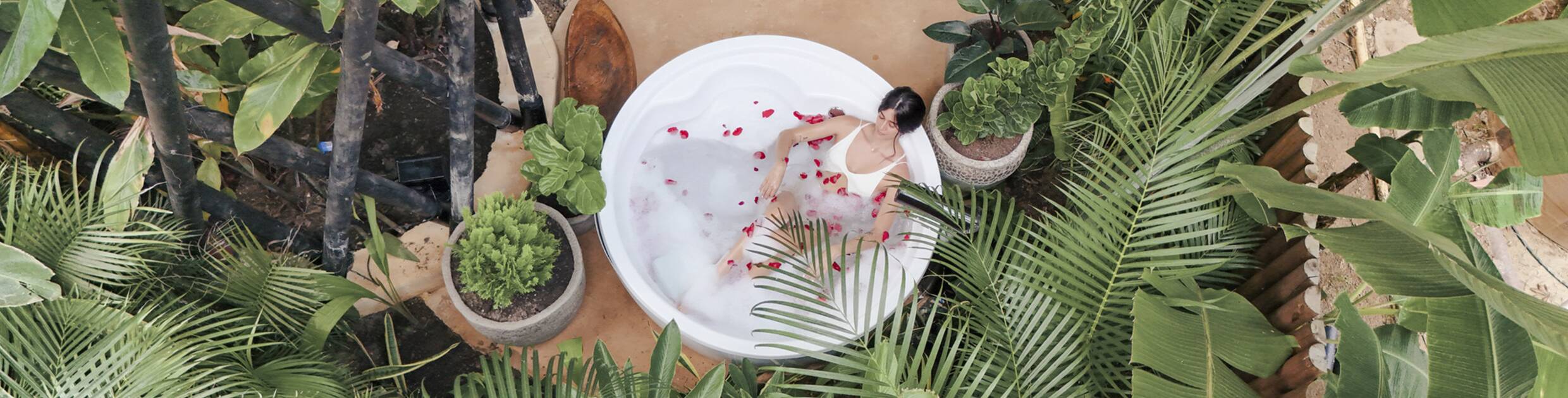Woman in a white swimsuit soaking in a round white tub with rose petals, surrounded by tropical plants.