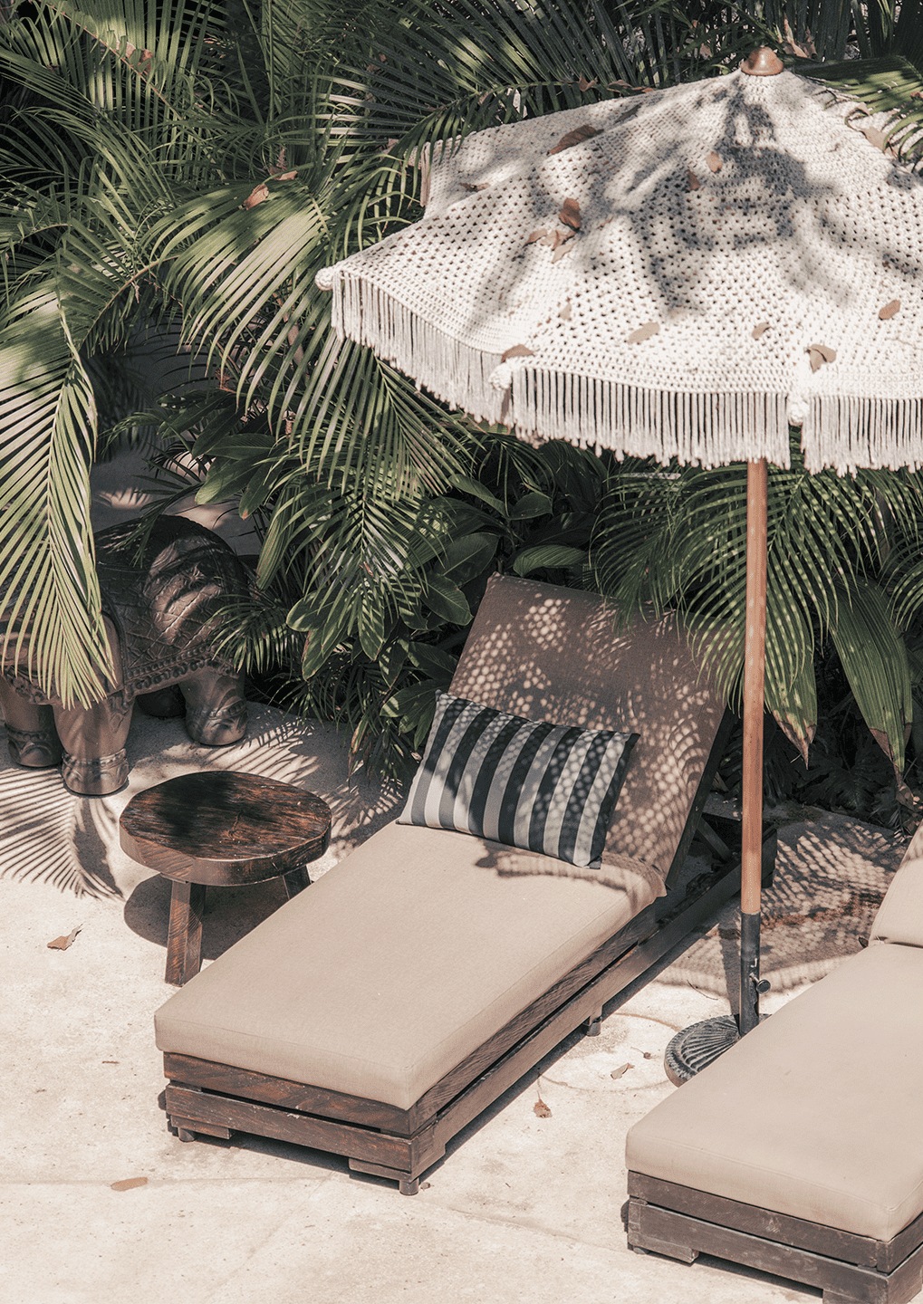 B1 1 - Casa Candela - Casa Candela Outdoor tropical seating area with a beige cushioned lounge chair, striped pillow, round wooden table, and a fringed parasol among palm leaves.