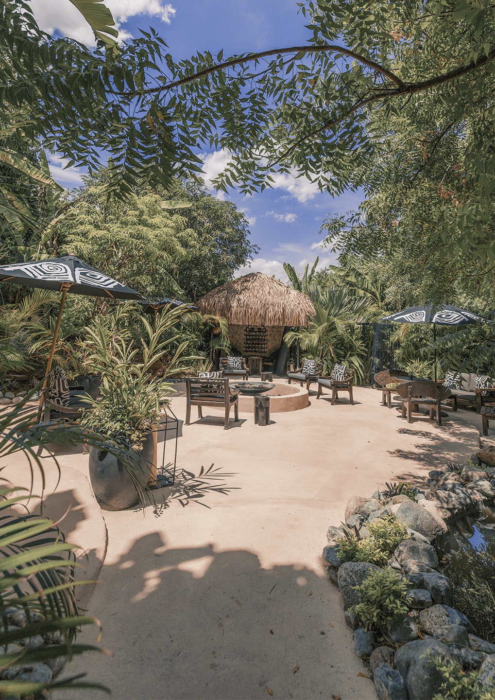 B2 - Casa Candela - Casa Candela Outdoor tropical lounge with a thatched hut, circular fire pit, and wicker chairs under patterned umbrellas.